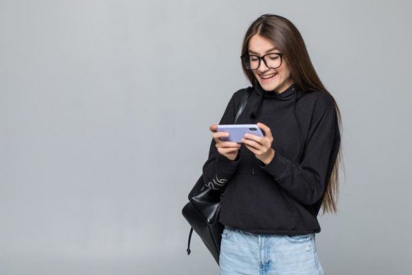 cheerful-young-girl-with-backpack-play-game-with-mobile-phone-isolated-white-wall_231208-1138